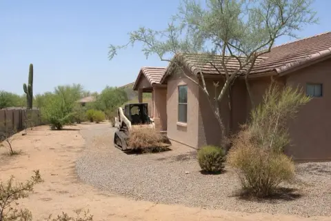 Desert-edge home with dried brush cleared in a wide radius, thinned native trees limbed up, and a gravel buffer along the stucco wall